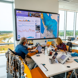 Team members working in a modern command center with laptops and tablets, analyzing environmental data displayed on a large wall screen showing charts and a coastal map.