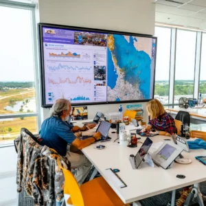 Team members working in a modern command center with laptops and tablets, analyzing environmental data displayed on a large wall screen showing charts and a coastal map.